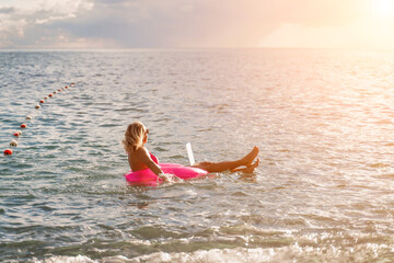 Woman works on laptop in sea. Freelancer, blond woman in sunglases floating on an inflatable big pink donut with a laptop in the sea at sunset. People summer vacation rest lifestyle concept.