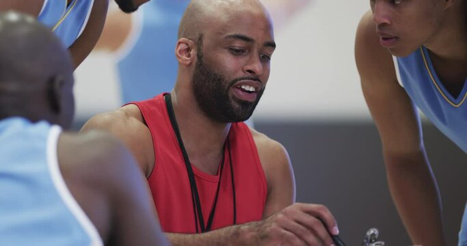 Diverse male basketball team and coach with clipboard in discussion at indoor court, in slow motion