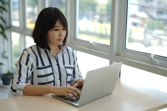 An Asian woman is working on a laptop seriously for business jobs in a cozy room.