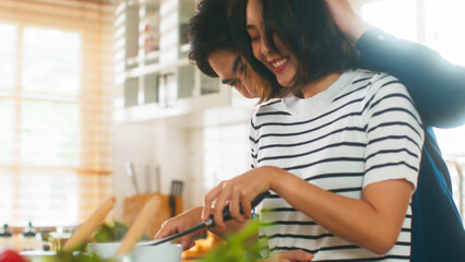 Attractive young Asian lover enjoy cooking healthy food having fun together. Handsome husband walk into camera romantic hug wife from behind. Beautiful couple making soup in the cozy kitchen at home