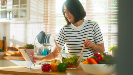 Beautiful young Asian woman hand finger slide tablet screen following healthy vegetarian food recipe in online cooking course. Attractive housewife mixing salad bell pepper, tomato, lettuce together - obrazy, fototapety, plakaty