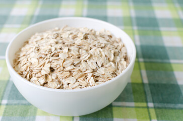 Raw oatmeal in a white bowl on a green checkered tablecloth. Concept of healthy food. Vegetarian and vegan. Horizontal orientation. Selective focus. Copy space.