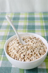 Raw oatmeal in a white bowl on a green checkered tablecloth with a teaspoon. Concept of healthy food. Vegetarian and vegan. Vertical orientation. Selective focus.