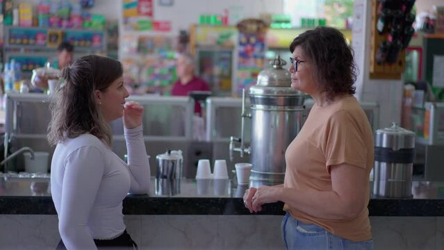 Two women talking at deli cafeteria counter, candid interaction. Customers chatting inside small business restaurant
