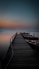 Wooden dock jutting out into a tranquil lake, with a variety of boats moored at its edges at sunset