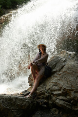 A woman sits on a rock and looks at a waterfall in the forest, a woman in a hat, a vacation in nature