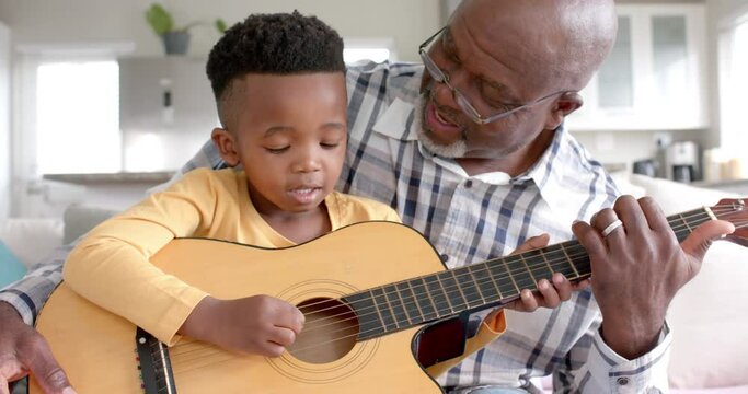 Happy african american grandfather and grandson playing guitar together at home, slow motion