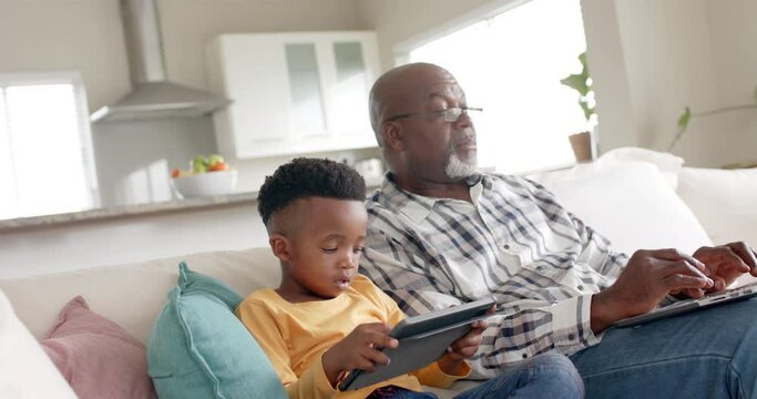 African american grandfather with grandson using tablet and laptop on couch at home, slow motion