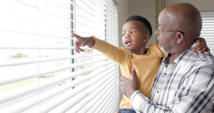 Happy african american grandson and grandfather embracing by window at home, slow motion