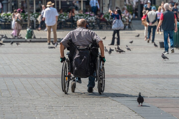 Authentic disabled person in a wheelchair on a sunny day in the city square among healthy people and pigeons. Integration, inclusion of people with disabilities into society