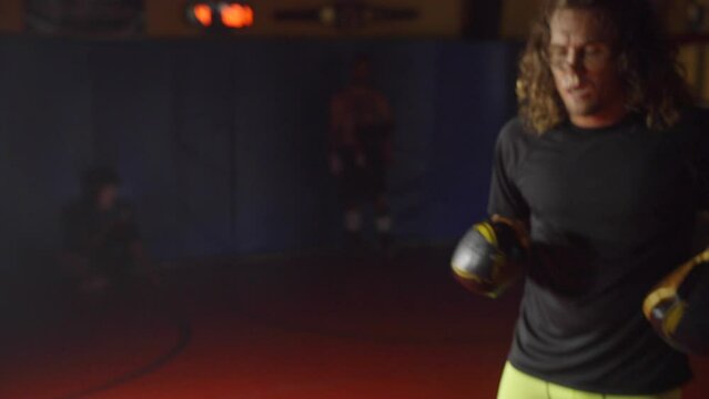 Male with long hair and a bold boxer with gloves fighting in training at a gym with sunlight