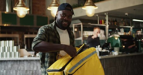 A food delivery man in a plaid shirt and black cap poses and smiles at a doner market. A man opens a special thermal bag for delivery