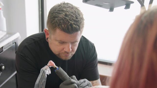Closeup of an artist man making a tattoo on a red hair female arm with glasses at a salon