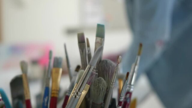 Pots of artists brushes in shallow depth of field, behind an artist works in home studio out of focus. Natural light, close up, daytime.