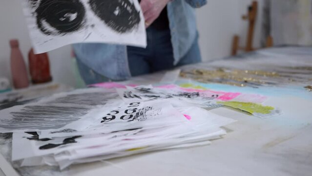 Female artist selecting swashes for canvas, stood in a home studio, surrounded by her paintings. Medium shot, daytime, natural light