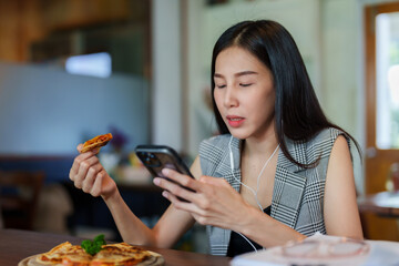 Asian woman sitting working in cafe, With relaxation, take break from work listen music by using headphones connected phone, picked up pizza from tray ate it deliciously, happy face when eating.