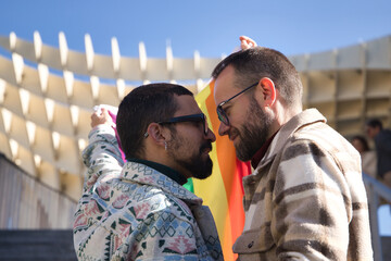 Real marriage of gay couple, looking at each other while holding a gay pride flag, happy and...