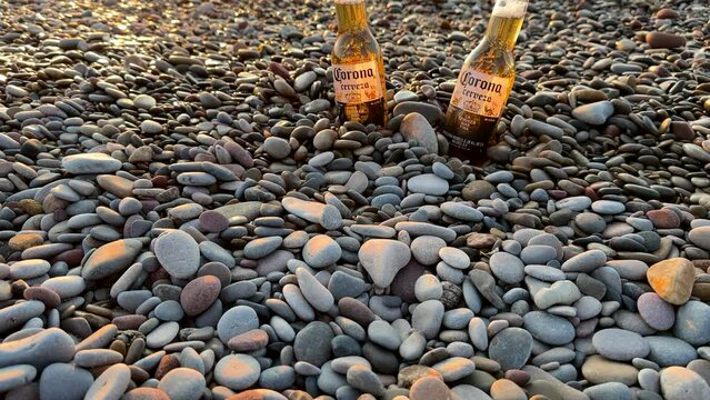 Beer Bottle on sunrise. Corona Cerveza Beer Bottle on Pebble stone beach on sunset sea. Сold beer on stones at sea beach. Corona Extra Coronita Mexican Lager Bottle. May 14, 2023, Spain, Valencia