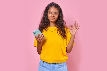 Young casual curly Indian millennial woman with smartphone shows OK gesture answering question how are or demonstrating agreement with you about quality of mobile phone stands in pink studio.