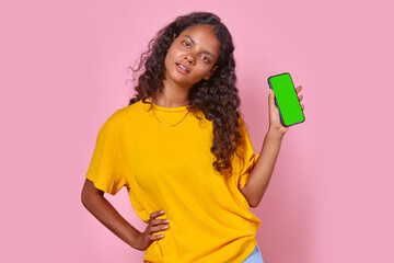 Young optimistic casual Indian woman teenager demonstrates green screen of phone and holds hand on belt offering to try new application for schoolchildren and students stands on pink background.