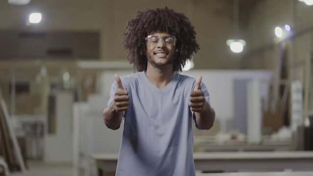 jovem Brasileiro de cabelo black power usando oculos de protecao sorrindo para a camera e fazendo sinal de positivo em uma fabrica no Brasil