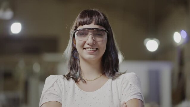 young student Brazilian woman with straight hair and bangs wearing safety glasses smiling at the camera and crossing her arms in a factory in Brazil