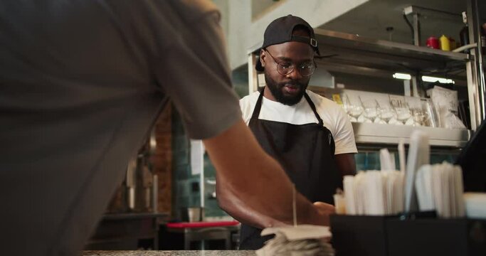 A Black person in a cap works in a doner market. A man with glasses takes an order and makes a check at a doner market