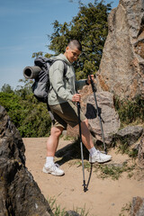 Young short haired female hiker with backpack holding trekking poles and looking at path while standing between stones with blue sky at background, trekking through rugged terrain, summer