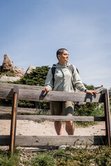 Fototapeta premium Smiling young short haired female traveler with backpack in casual clothes looking away while standing near wooden fence with nature at background, hiker trekking through landscape, summer