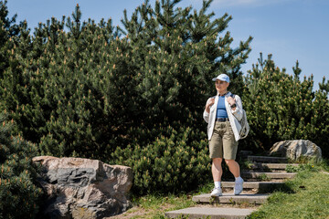 Naklejka na ściany i meble Young female hiker in casual clothes and baseball cap holding backpack while walking down on stairs with nature and blue sky at background, curious hiker exploring new landscapes