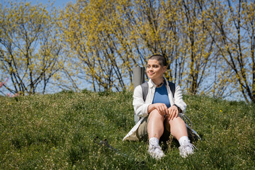 Smiling short haired young female hiker with backpack and fitness mat looking away while sitting and resting on grassy hill and resting, explorer woman discovering hidden trails