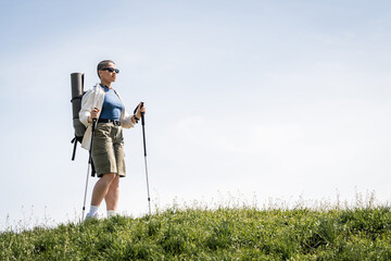 Short haired young female traveler in sunglasses with backpack and travel equipment holding trekking poles while walking on grassy hill , explorer woman discovering hidden trails