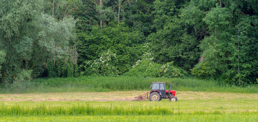 Zgrabianie siana za pomocą zgrabiarki przyłączonej do czerwonego traktora, na łące u stóp wzgórza porośniętego lasem liściastym . - obrazy, fototapety, plakaty
