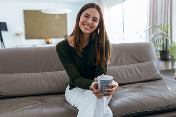 Beautiful young woman drinking a cup of coffee while sitting on couch in living room at home.
