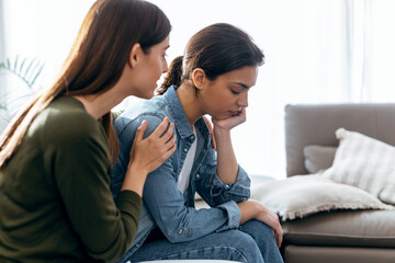 Pretty young woman supporting and comforting her sad friend while sitting on the sofa at home.