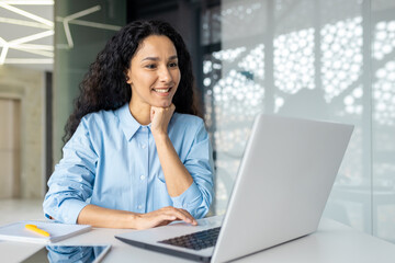 Happy and smiling hispanic businesswoman typing on laptop, office worker with curly hair ,happy with achievement results, at work inside office building