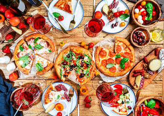 Fototapeta na wymiar Pizza party table. Top view glasses with red wine and dinner plates on rustic wooden table with hot pizzas, italian appetizers, salads, cheese, fruits and berries. Family lunch with fast food