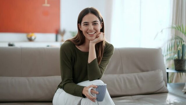 Video of beautiful young woman drinking a cup of coffee while sitting on couch in living room at home.