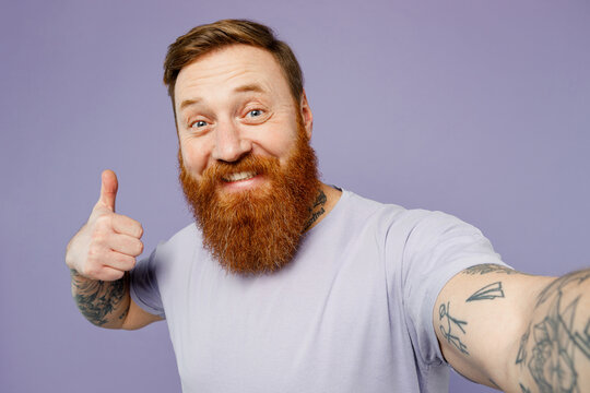 Close up young redhead bearded man he wear violet t-shirt casual clothes doing selfie shot pov on mobile cell phone show thumb up isolated on plain pastel light purple background. Lifestyle concept.