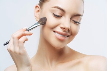 Positive European woman applies foundation with powder brush, has healthy skin, clean complexion, natural makeup, indoors