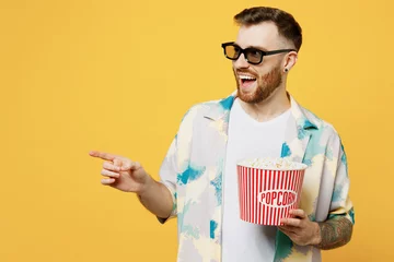 Fototapeten Young maan in 3d glasses wears blue shirt white t-shirt casual clothes watch movie film hold bucket of popcorn in cinema point index finger aside isolated on plain yellow background studio portrait. © ViDi Studio