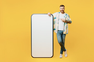 Full body fun young man he wear blue shirt white t-shirt casual clothes point index finger on big huge blank screen mobile cell phone smartphone with area isolated on plain yellow background studio.