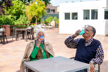 In a vibrant garden of a senior care home, a happy elderly couple savor their drinks, cherishing...