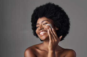 Beauty, skin care and black woman with lotion on face, afro and cosmetics in studio on grey...