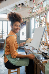 African American mature female artist smiling at camera while painting on easel in art studio
