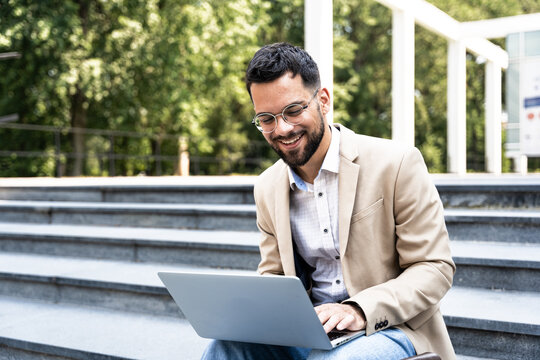 Handsome Man Working On Laptop In City Street. Man Using His Knowledge While Sitting On Stairs Outdoors. Businessman Successful Finishing Task Project On Computer In-front Of Office Building