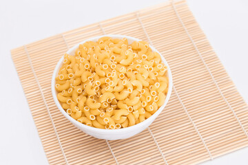 Uncooked Chifferi Rigati Pasta on White Cup on Bamboo Mat on White Background. Fat and Unhealthy Food. Scattered Classic Dry Macaroni. Italian Culture and Cuisine. Raw Pasta