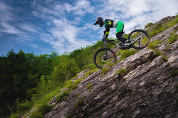 Downhill rider cyclist rides down a steep cliff surrounded by green trees against a blue sky. Copy space mtb concept