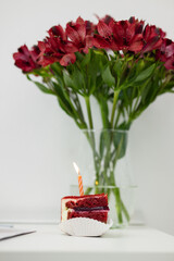 A red slice of cake with a candle on a white table. Bouquet of red flowers