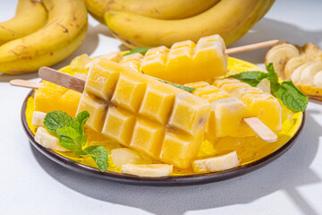 Bright yellow vegan banana ice cream popsicles. Frozen banana fruit lollypops, with ice and mint decor, white kitchen background. Summer cold vegetarian healthy dessert, copy space
